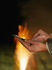 Cropped image of hand using smart phone against campfire burning at night
