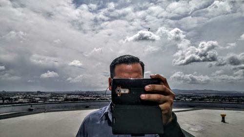 Man photographing with mobile phone while standing on building terrace against cloudy sky