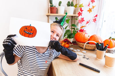 A cheerful girl in a witch costume shows her drawing on halloween, looking out from behind it.