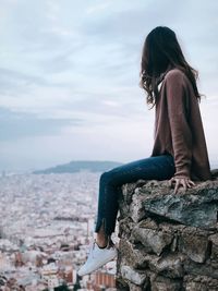 Side view of woman looking at city while sitting on cliff against sky