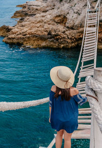 Rear view of young woman standing on old rope bridge over sea cove