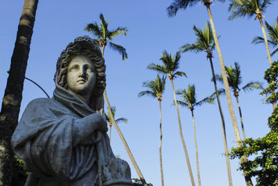 Low angle view of statue by coconut palm trees against clear blue sky