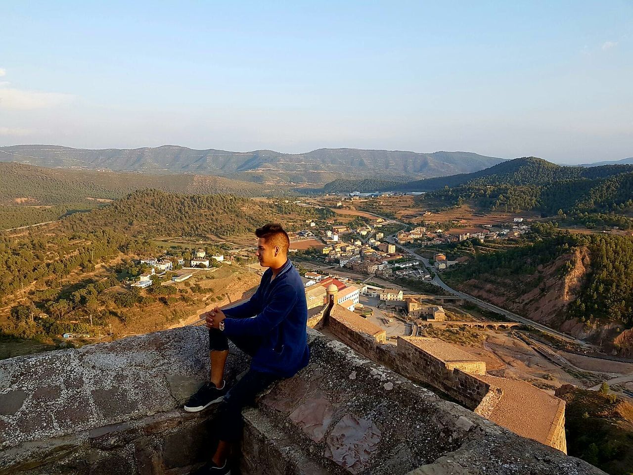 PORTRAIT OF YOUNG MAN SITTING ON RETAINING WALL AGAINST MOUNTAIN