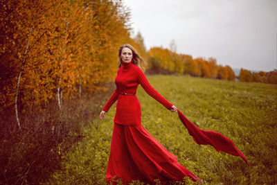 Woman in a red dress and sweater stands in a field with trees in autumn