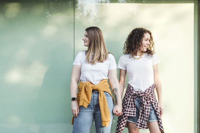 Smiling beautiful women holding hands while looking away against wall