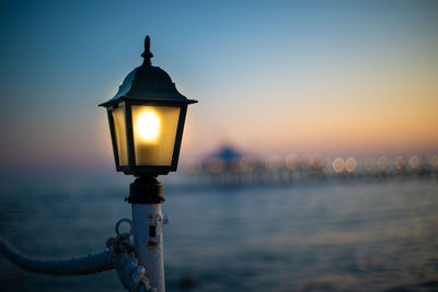 Illuminated street light by sea against sky during sunset