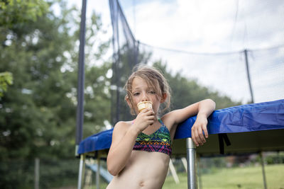 Low angle portrait of girl eating ice cream while standing against trampoline