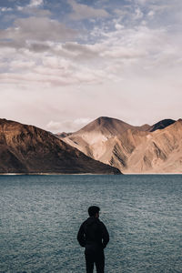 Rear view of silhouette man standing at lakeshore against mountains