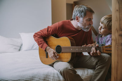 Father playing guitar in front of son while sitting on bed at home