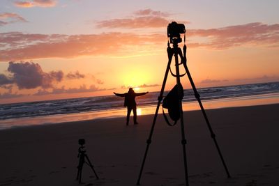 Silhouette man and tripod on beach against sky during sunset