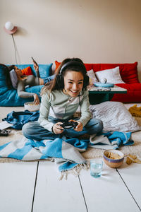 Young woman playing video game while friend using smart phone on sofa at home