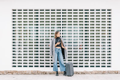 Female tourist with luggage standing alone on the street and browsing mobile phone while getting lost in city looking away