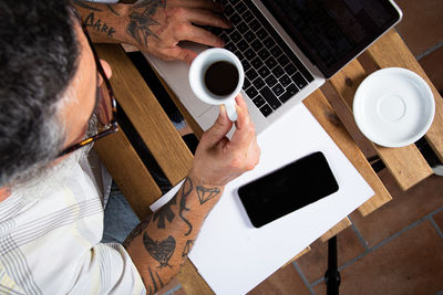 From above of crop unrecognizable male in casual clothes with tattooed arms drinking coffee and browsing laptop while working remotely at table in home workplace