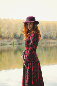 Portrait of young redhead woman standing by lake against sky