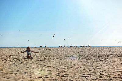 Girl with arms outstretched sitting at beach against clear sky during sunny day