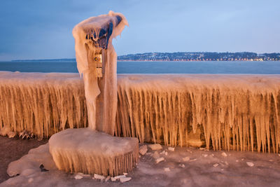 Frozen coin-operated binoculars and railing by lake against sky at dusk