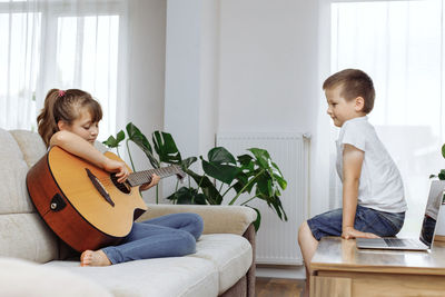 Brother and sister are resting at home. the girl sits on the couch and tries to play the guitar