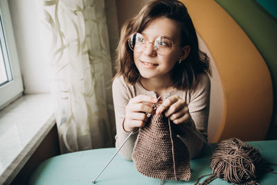Teenage girl in a beige sweater, wearing glasses knitting needles standing by the window. 