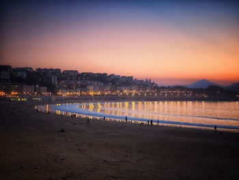 Illuminated city by sea against sky at sunset