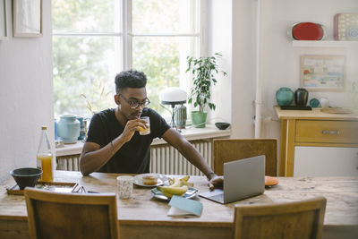 Young man drinking juice while looking at laptop at table in house