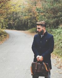 Thoughtful man with bag standing on road against trees
