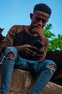 Midsection of young adult sitting using a mobile phone against blue sky