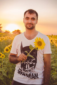Portrait of smiling man holding sunflower while standing on field during sunset