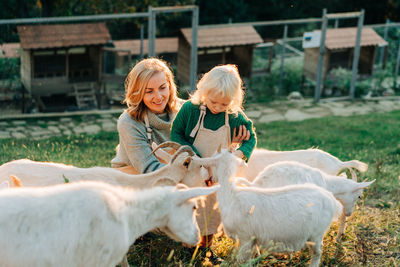 Happy smiling mom and little daughter feed and care for goats on the farm. 