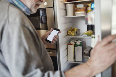 Senior man holding phone while searching in refrigerator at home