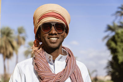 Cheerful arab male in sunglasses wearing traditional turban looking at camera and smiling while standing on blurred background of exotic city in summer