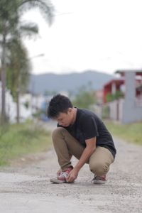 Young man tying shoelace while crouching on footpath against sky