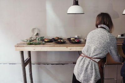 Rear view of female entrepreneur standing by table with food plates against wall at workshop