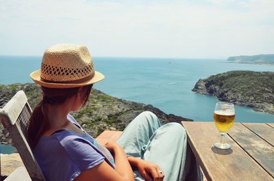 Woman sitting with hat by beer against sea and sky on sunny day