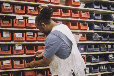 Blue-collar worker searching in containers on rack while working in workshop