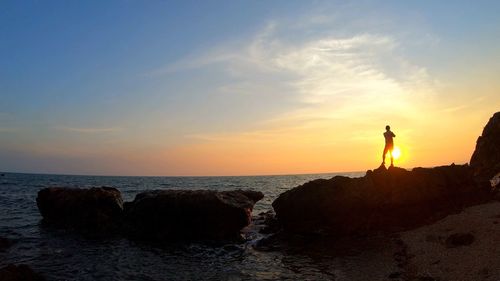 Man standing on rock by sea against sky during sunset