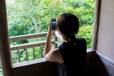 Woman photographing through mobile phone while standing by window