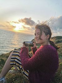 Side view of woman photographing sea against sky during sunset