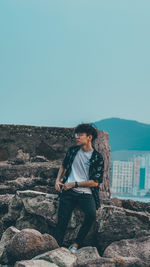 Young man sitting on rock against clear sky