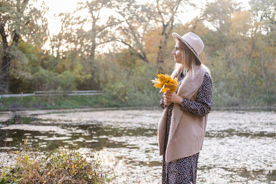 Beautiful young stylish woman in hat holds autumn leaves, walks in nature in autumn park in fall