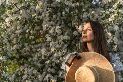 Dreamy brunette woman with straw hat enjoys the atmosphere in spring blooming apple tree garden.