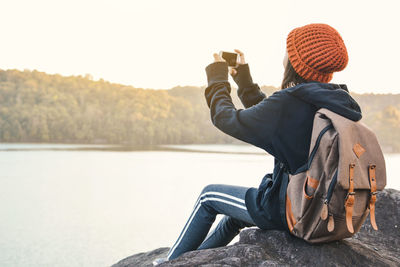 Girl photographing through mobile phone while sitting against lake and sky