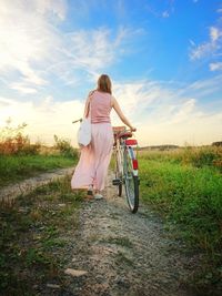 Rear view of woman with bicycle walking on land against sky during sunset