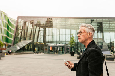 Side view of senior male commuter looking away while holding smart phone against railroad station building