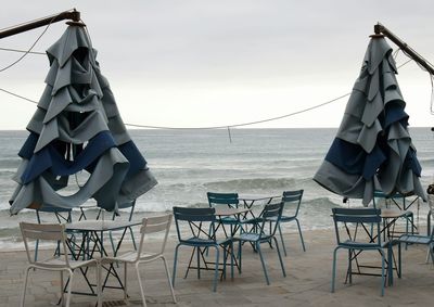 Empty chairs and tables at beach against sky