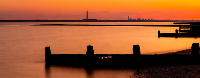 Silhouette wooden posts in sea against orange sky