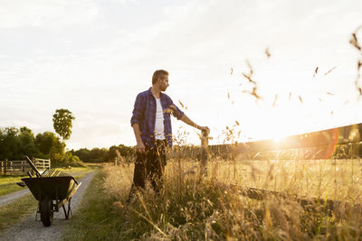 Farmer standing by fence and wheelbarrow on grassy field during sunny day