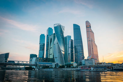 Modern buildings by river against sky during sunset