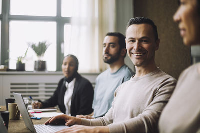 Happy mature businessman with laptop sitting by colleagues in meeting room