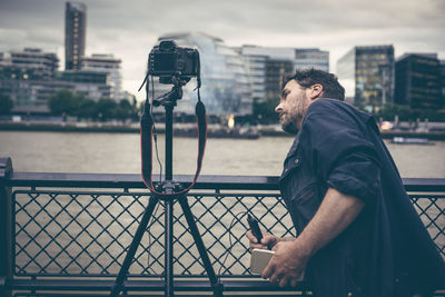 Photographer adjusting camera at railing by river in city