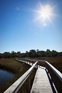 Empty boardwalk by field against sky on sunny day